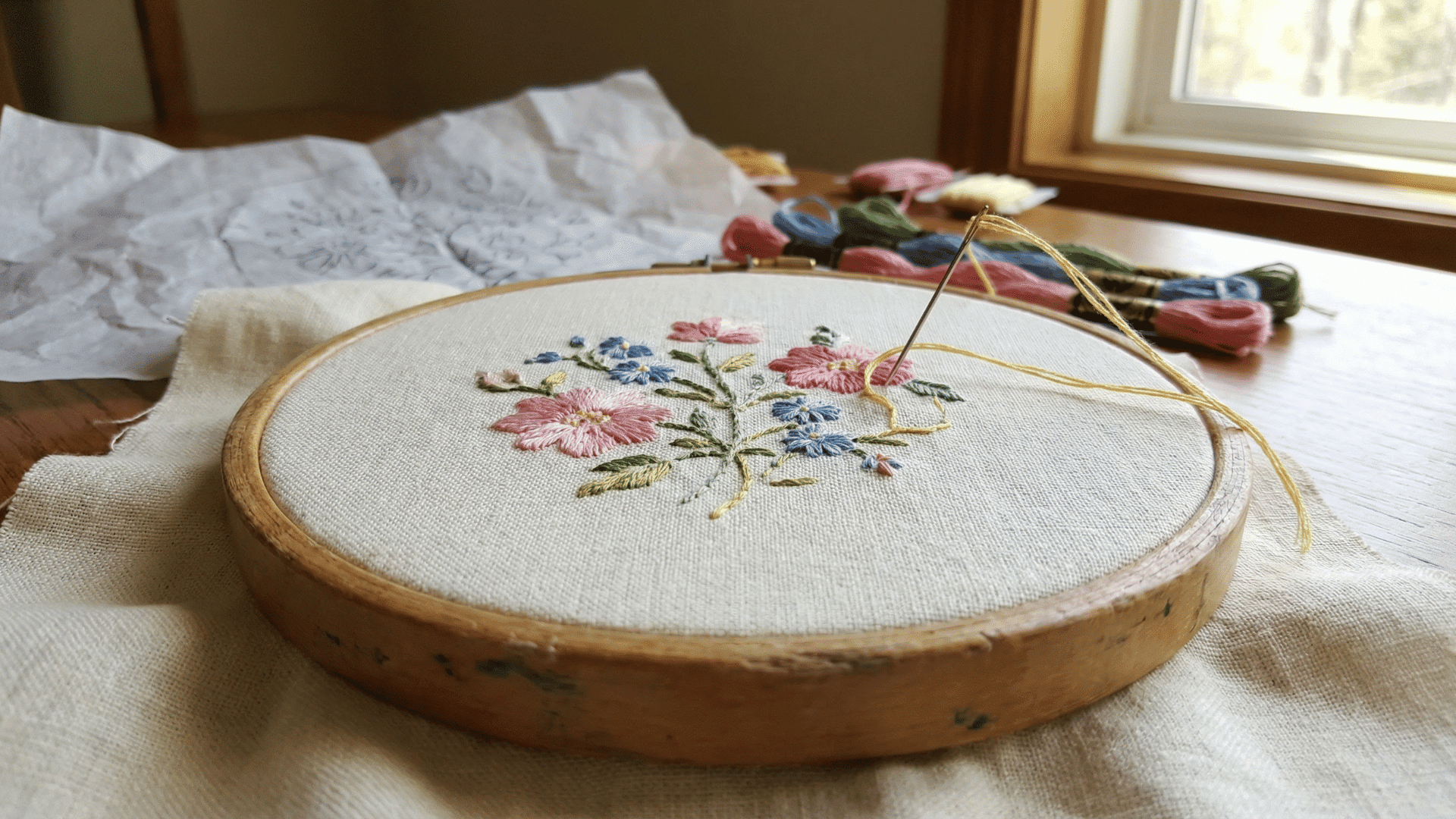 a floral embroidery design featuring pink and blue flowers in a wooden hoop, shown with a needle, thread, and pattern paper.