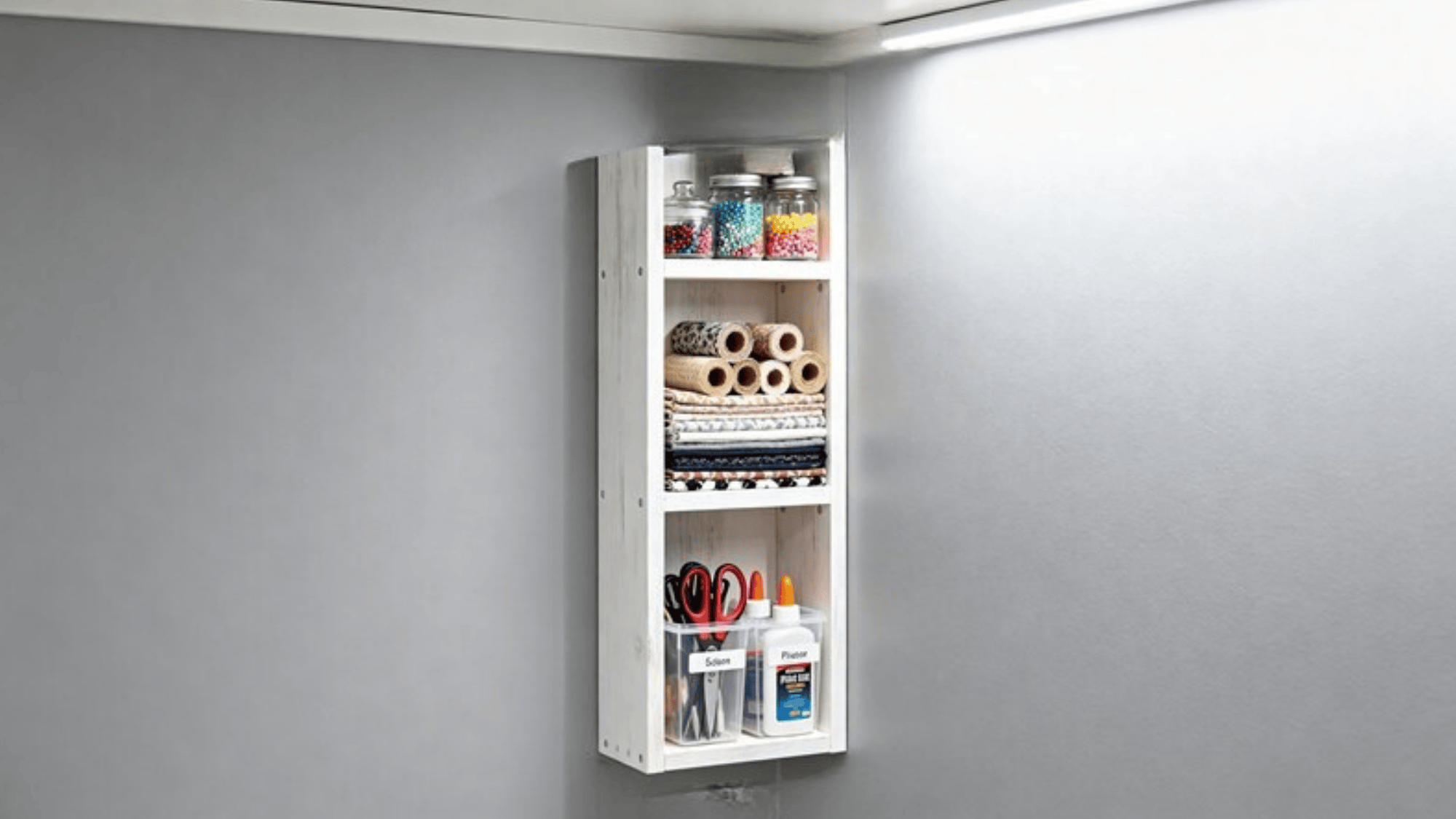 a narrow white storage shelf mounted on a wall, filled with craft supplies like beads, fabric rolls, scissors, glue, and a small potted plant, with measurements marked on the side