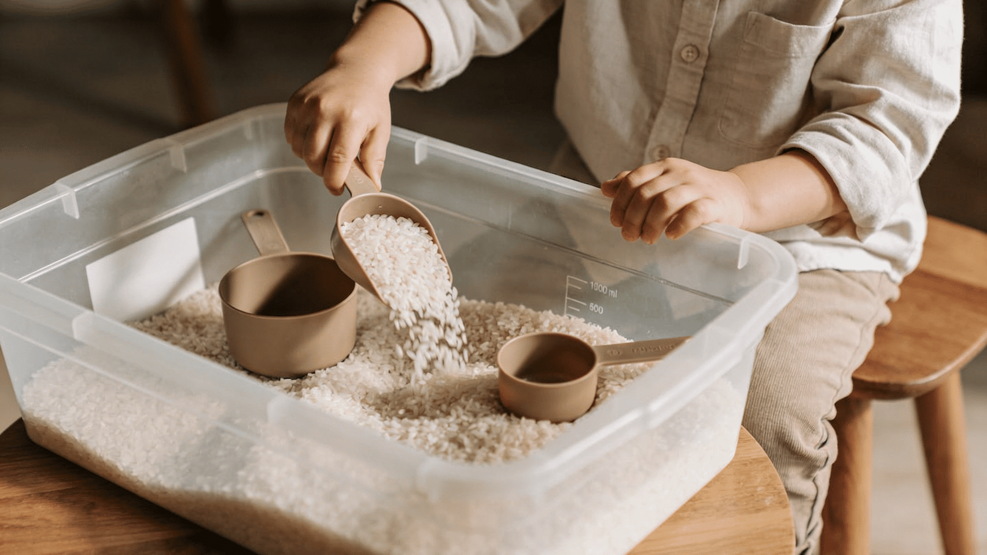 a preschool child uses a tan scoop to pour white rice into a measuring cup inside a clear plastic sensory bin.