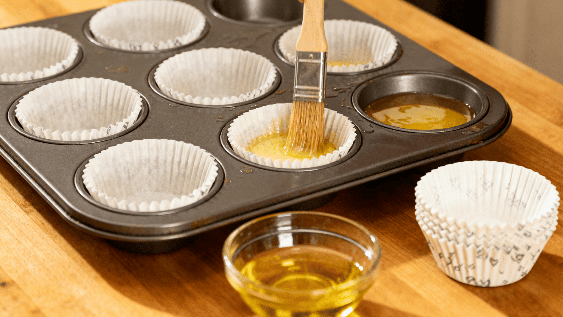 brushing oil inside a muffin tin lined with paper cups preparing the tray before adding muffin batter