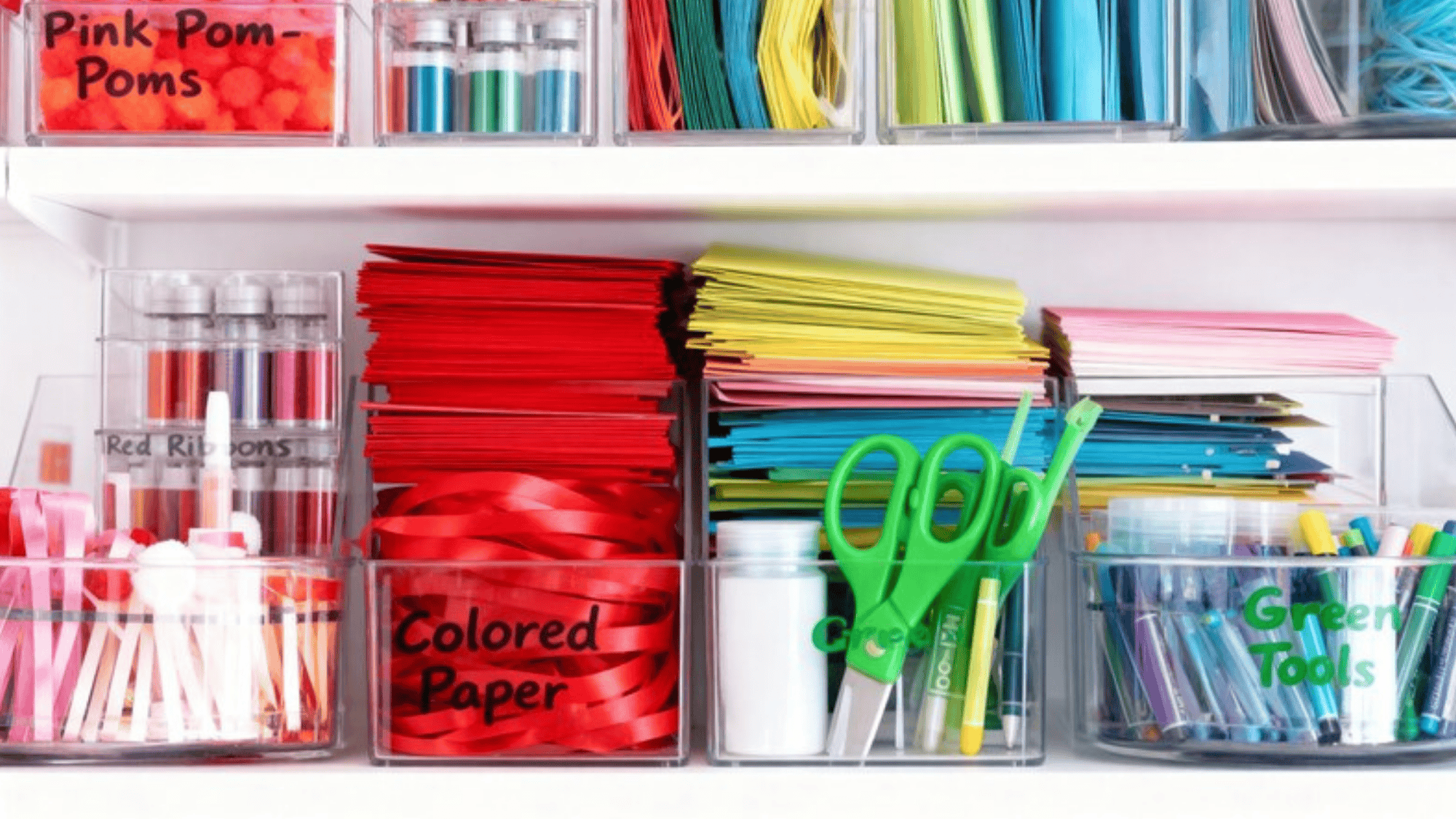 craft supplies on a shelf, including colored paper, ribbons, pom-poms, scissors, and markers, all neatly labeled and stored in clear bins