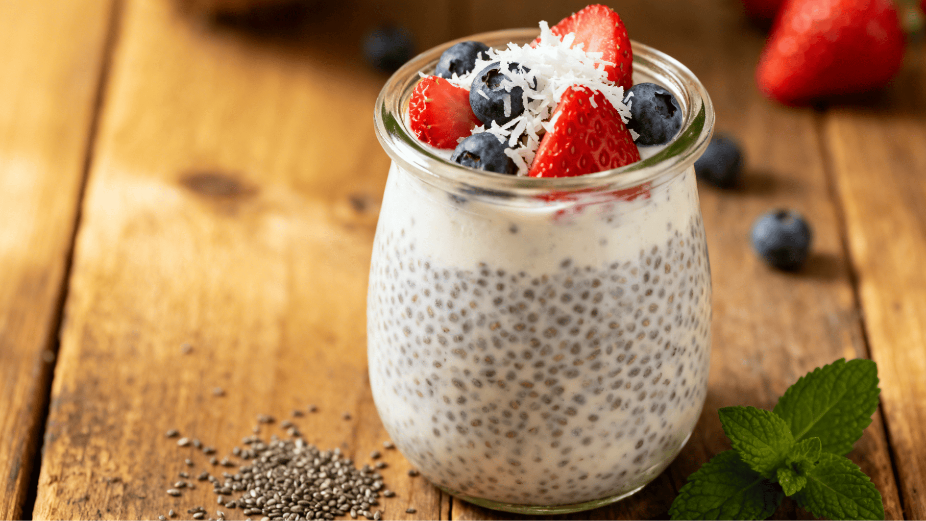 glass jar of coconut chia pudding topped with strawberries blueberries and coconut flakes placed on a wooden table with mint leaves nearby