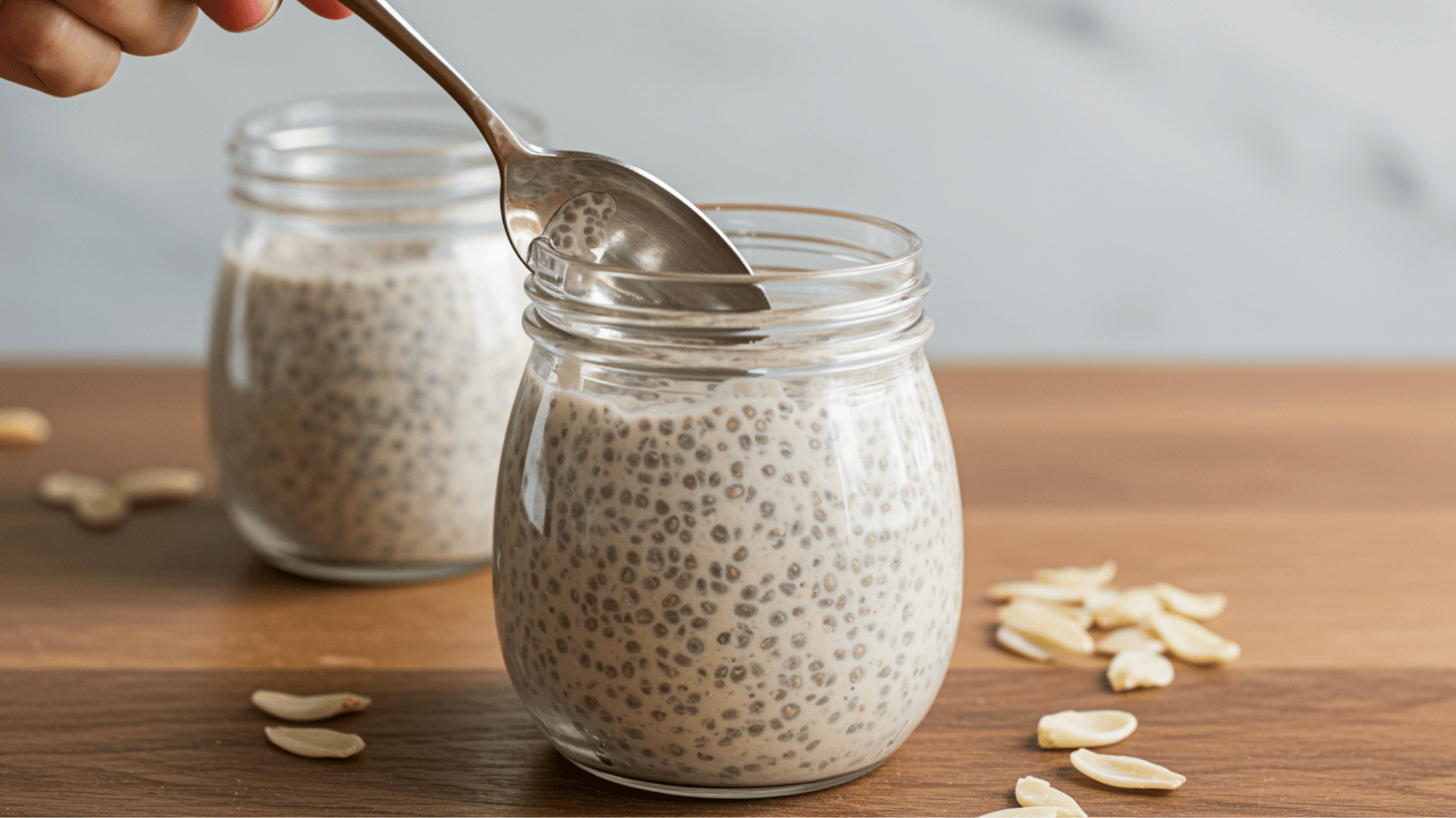 hand holding a spoon above a jar of coconut chia pudding showing its thick texture with almond slices scattered on the table