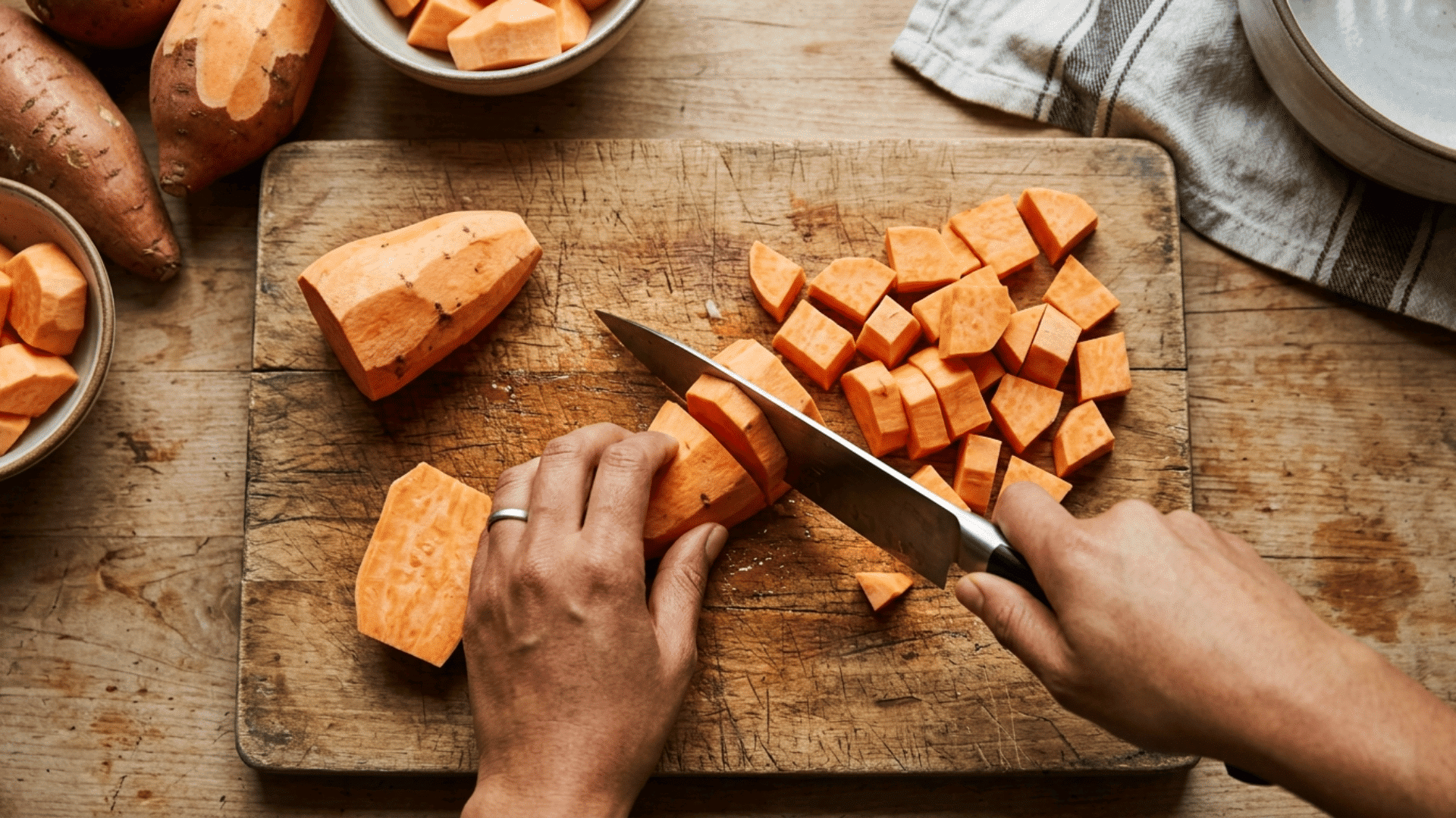 hands slicing fresh sweet potatoes on a wooden board in a bright kitchen.