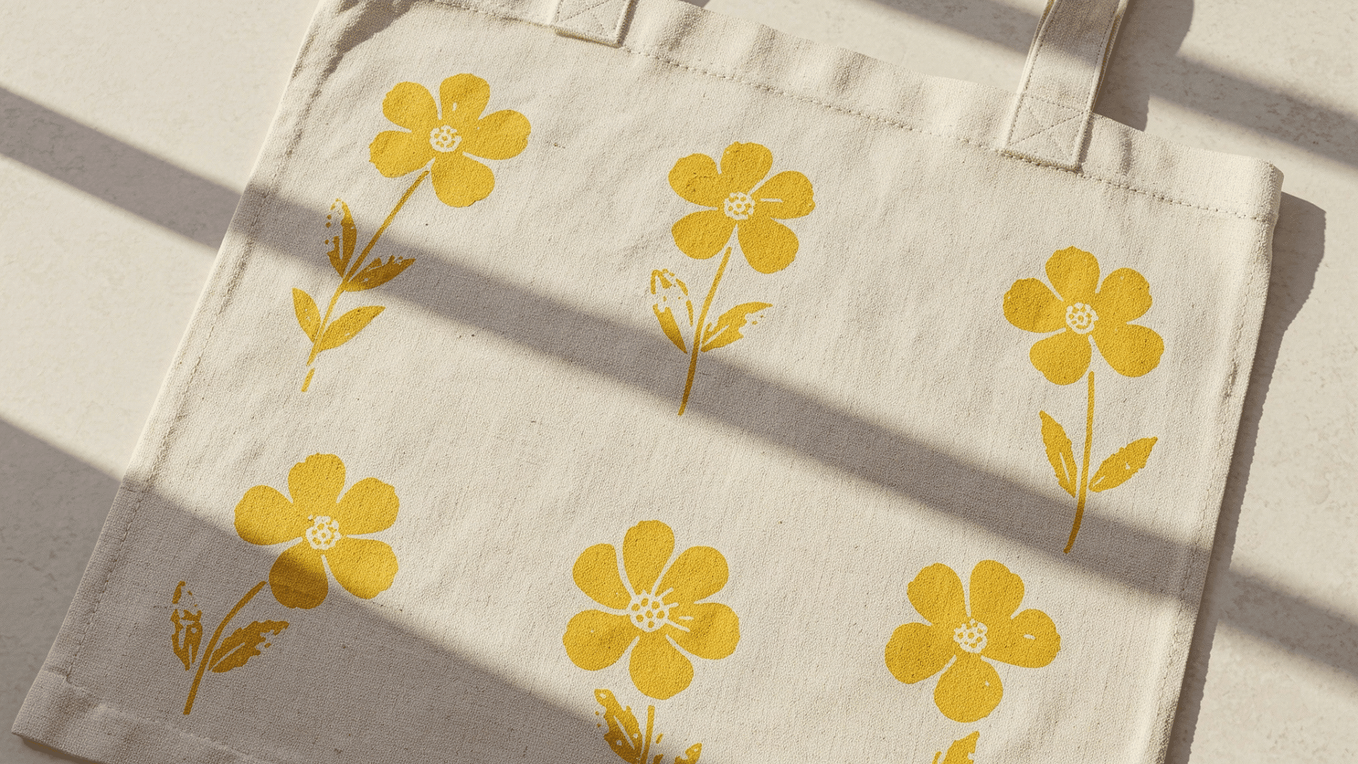 off-white canvas tote bag featuring a simple hand-stamped pattern of six bright yellow flowers in a sunlit setting.