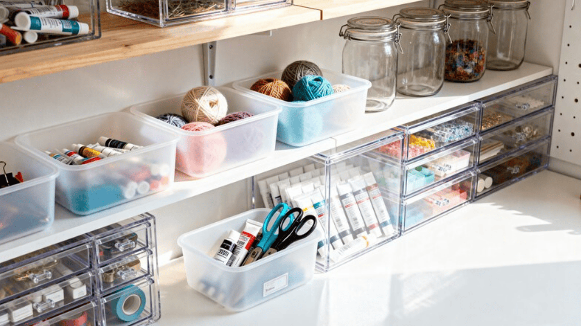organized craft supplies on a shelf, including yarn balls, paint tubes, scissors, and jars of colorful beads, neatly stored in clear plastic bins and drawers