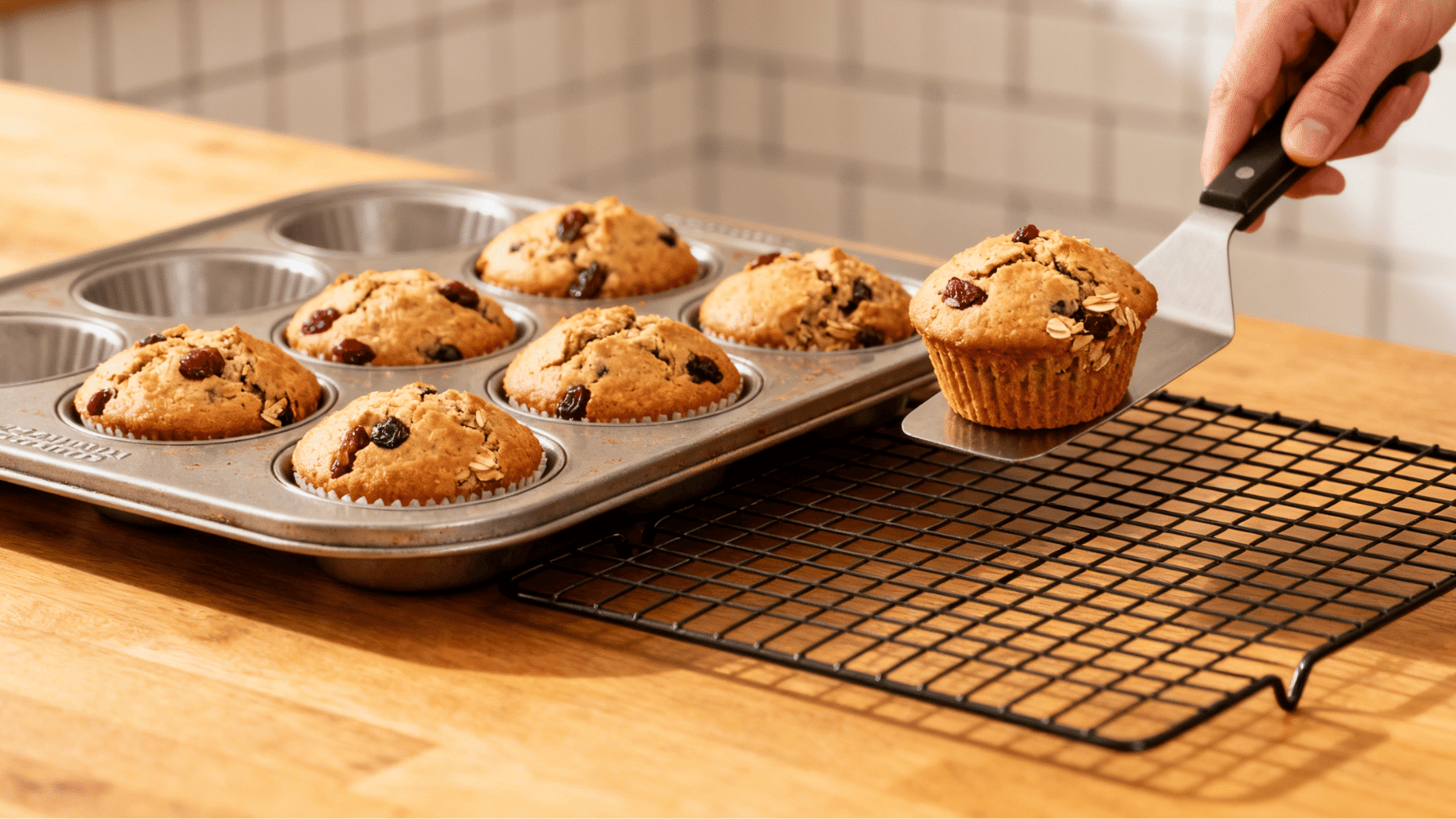 removing freshly baked raisin muffins from a muffin tray and placing one onto a cooling rack with a spatula