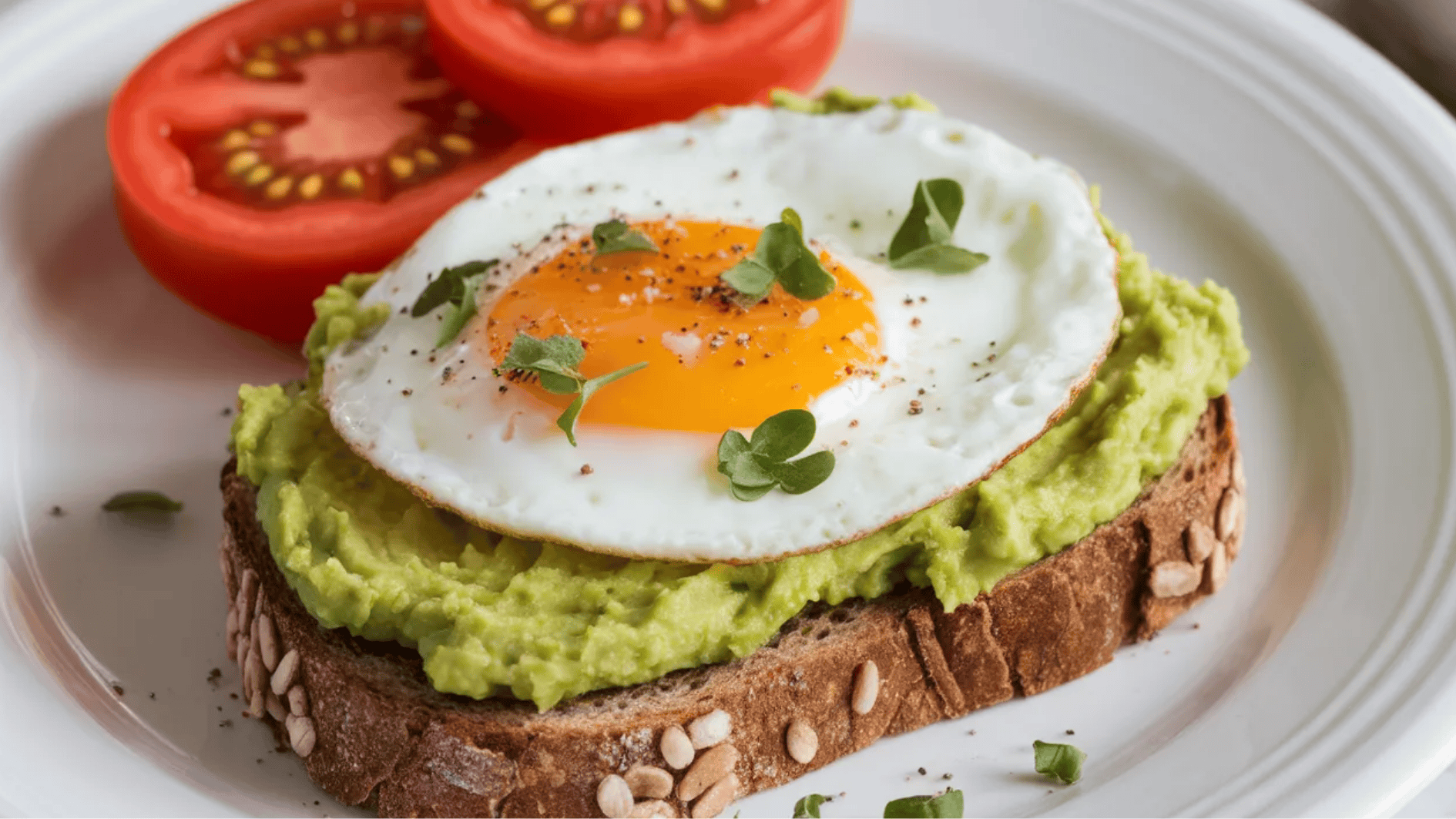 roasted bread topped with mashed avocado, a fried egg, fresh herbs, and a side of tomato slices