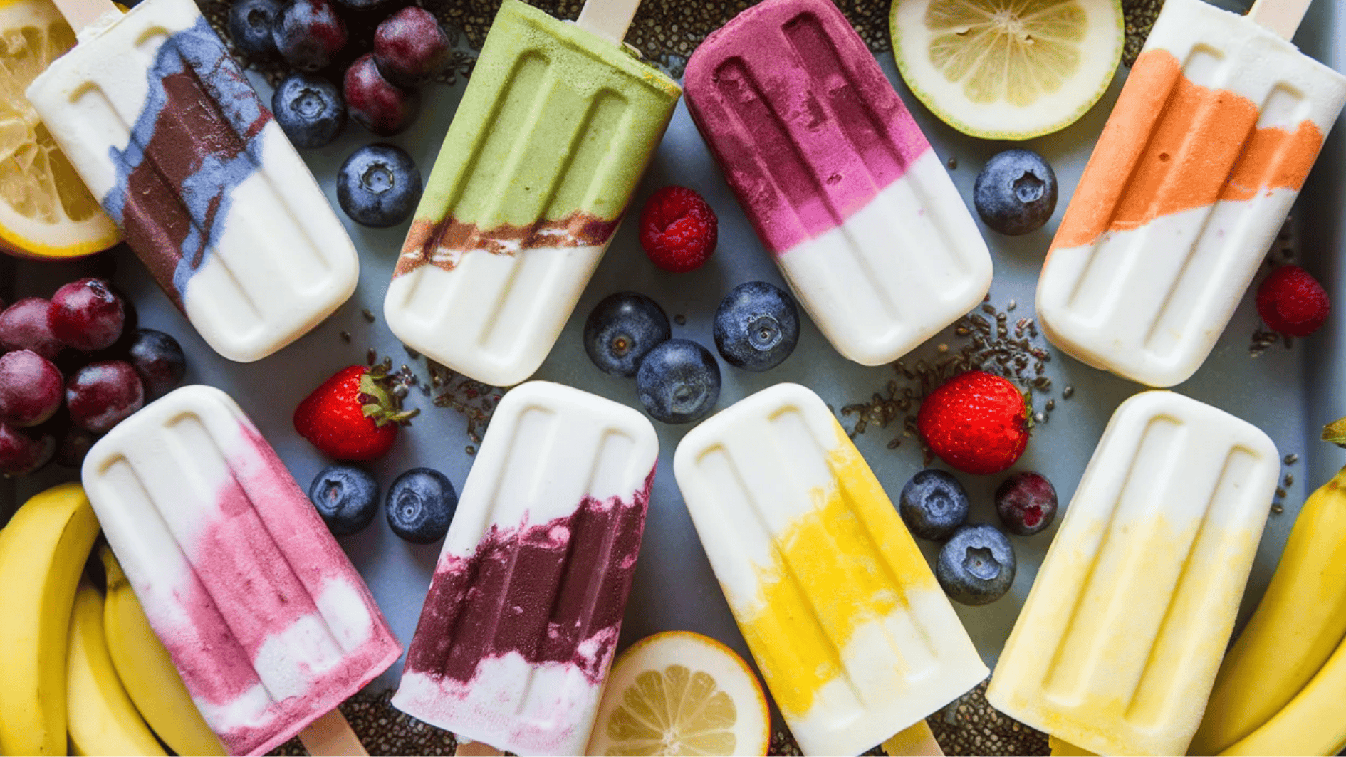 variety of colorful fruit popsicles with yogurt layers, surrounded by fresh fruit like strawberries, blueberries, and bananas