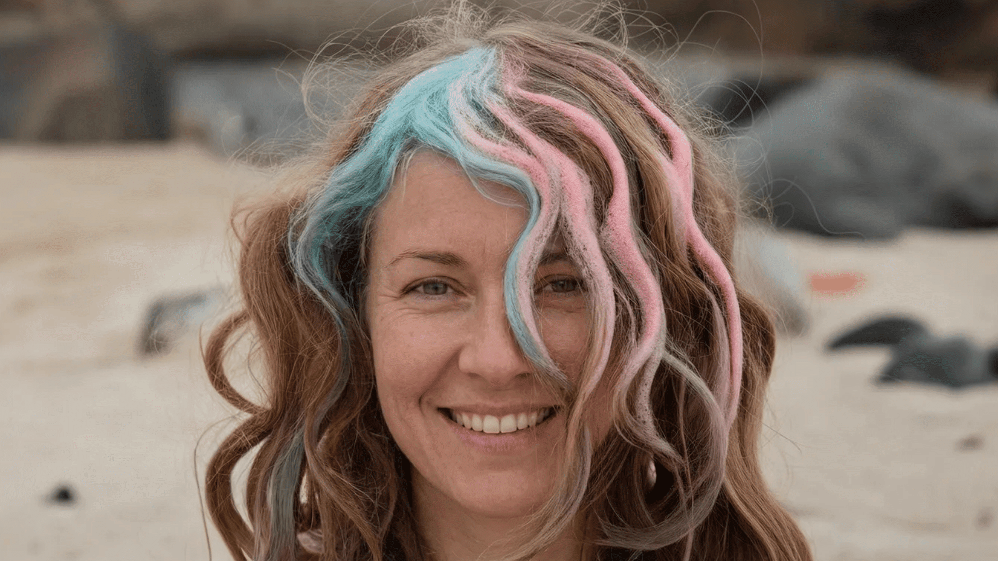 woman with long, curly hair featuring pastel pink and blue streaks, standing outdoors with a beach backdrop