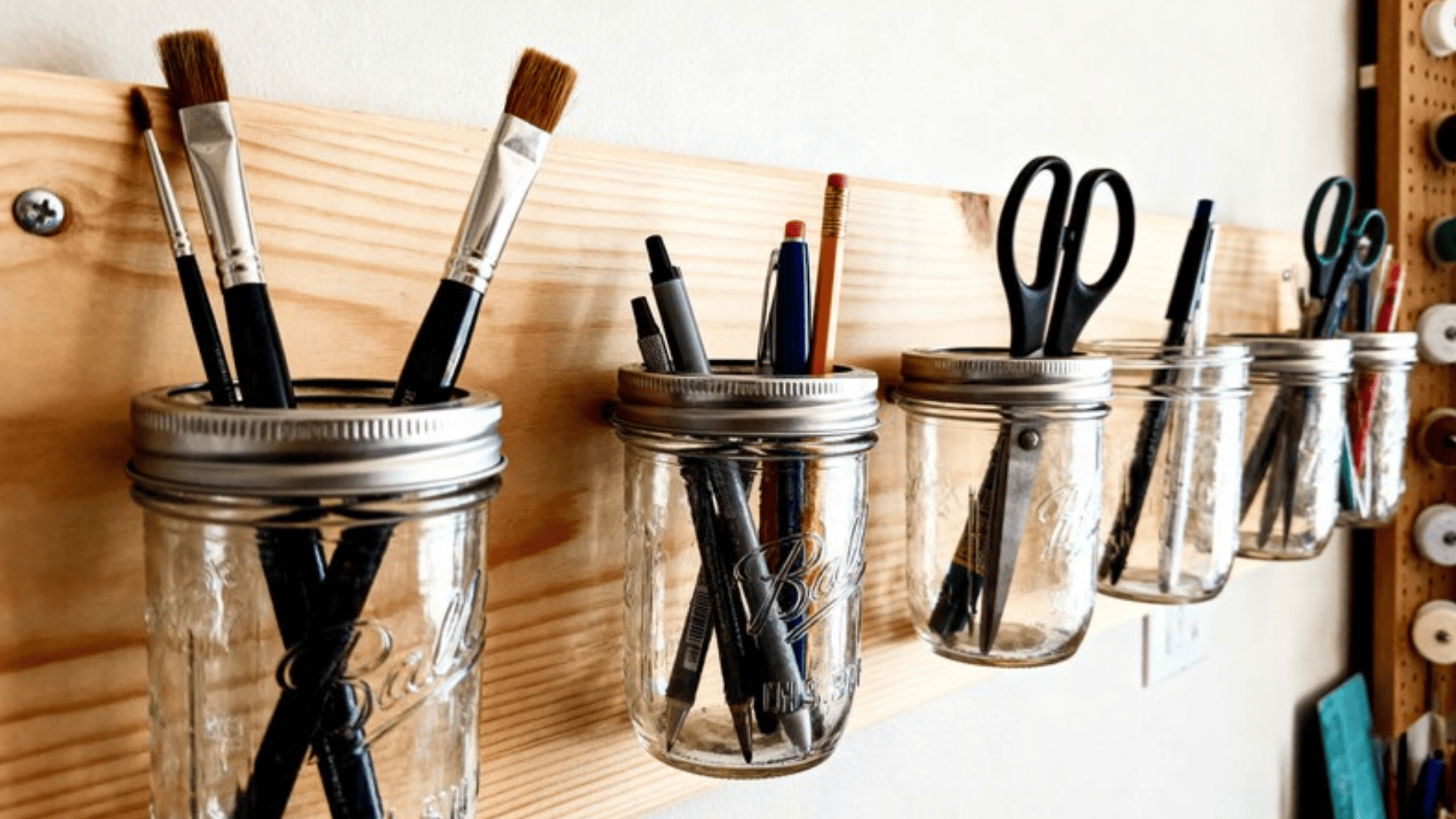 wooden wall-mounted shelf with mason jars holding brushes, pens, scissors, and pencils, creating an organized and functional workspace